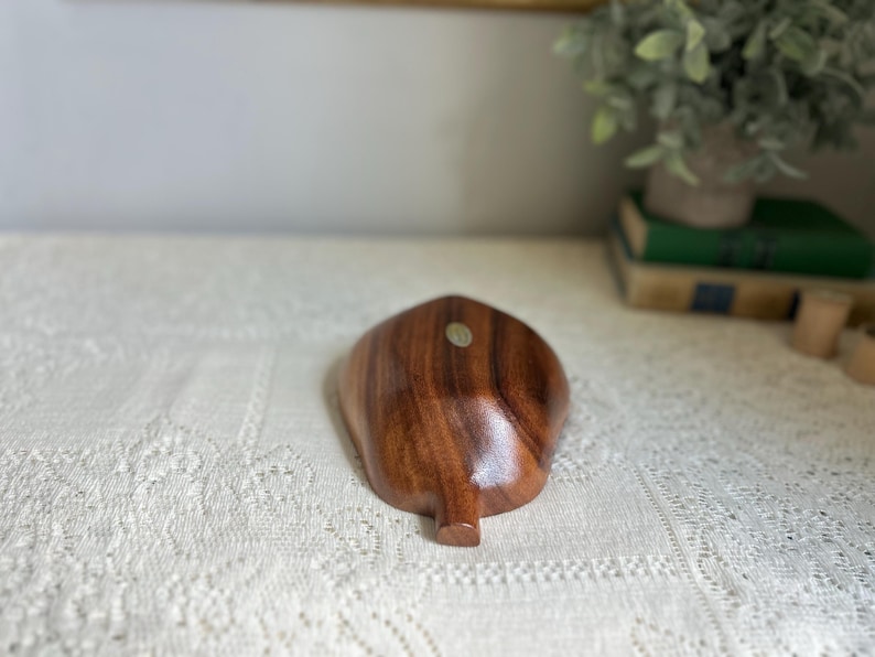 K&ouml;nnte beinhalten: Eine holzfarbene, blattf&ouml;rmige Schale mit dunkelbraunem Finish. Die Schale hat eine glatte, polierte Oberfl&auml;che und einen kleinen, abgerundeten Stiel. Ein kleiner Aufkleber ist auf der Schale sichtbar. Die Schale wird wahrscheinlich zu dekorativen Zwecken verwendet.
