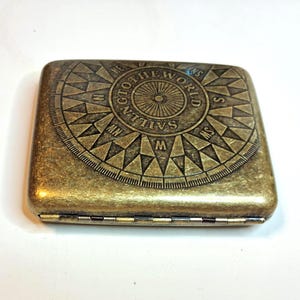 K&ouml;nnte beinhalten: Ein rechteckiges, antik-messingfarbenes Etui mit einem Kompassrosen-Design auf dem Deckel. Die Kompassrose zeigt Richtungsanzeigen und den Text "SAILING TO THE WORLD". Das Etui hat eine Klapp&ouml;ffnung.