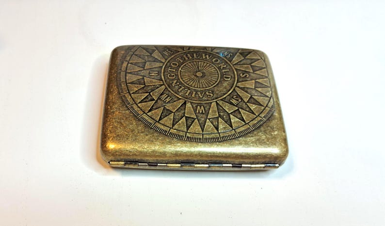 K&ouml;nnte beinhalten: Ein rechteckiges, antik-messingfarbenes Etui mit einem Kompassrosen-Design auf dem Deckel. Die Kompassrose zeigt Richtungsanzeigen und den Text "SAILING TO THE WORLD". Das Etui hat eine Klapp&ouml;ffnung.
