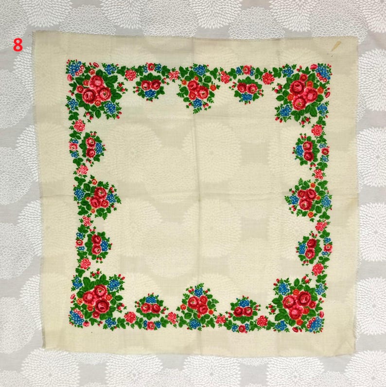 May include: Cream-colored square textile with a floral border. The border features clusters of red roses, blue flowers, and green leaves. The number "8" is visible in the upper left corner.