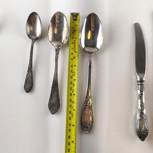 Op de afbeelding: Zilveren bestekset met sierlijke handvatten, waaronder lepels van verschillende maten en een mes. Een gele meetlint is naast de lepels geplaatst. De lepels zijn ongeveer 15 tot 20 cm lang.