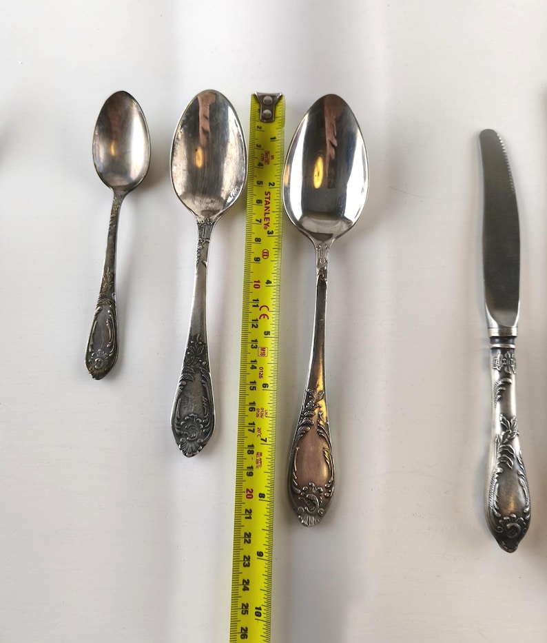 Op de afbeelding: Zilveren bestekset met sierlijke handvatten, waaronder lepels van verschillende maten en een mes. Een gele meetlint is naast de lepels geplaatst. De lepels zijn ongeveer 15 tot 20 cm lang.