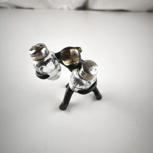 以下が含まれることがあります： ヴィンテージの塩コショウ入れセット。銀色の蓋が付いた2つの透明なガラスシェーカーが、金色の顔が付いた黒い金属製のスタンドで支えられています。スタンドには3つの脚があります。