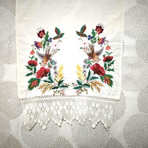 May include: White embroidered textile featuring a floral design with red, purple, and green flowers, and brown birds. The bottom edge has a white crocheted lace trim. The textile is likely a decorative piece.