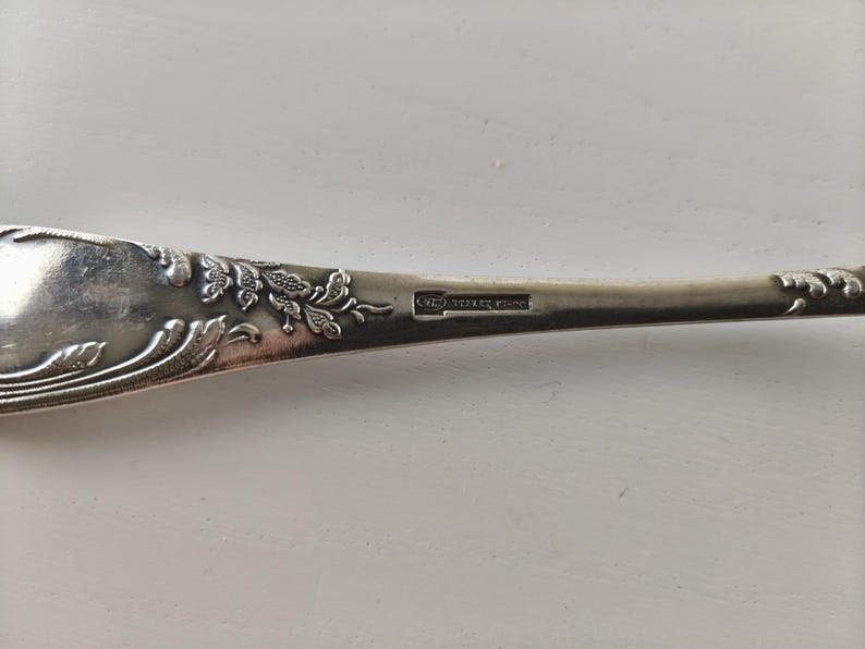 Op de afbeelding: Close-up van een zilveren lepel met sierlijke bloemendetails. De steel van de lepel heeft reli&euml;fbladeren en wijnranken, met een kleine rechthoekige markering. De lepel staat tegen een witte achtergrond.