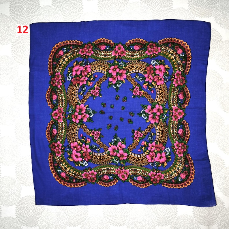 Op de afbeelding: Een vierkante, blauwe sjaal met een bloemenmotief. De sjaal heeft een centraal patroon van roze bloemen en groene bladeren, omlijst door een goudbruine rand. De randen zijn versierd met een geschulpte rand. Het nummer "12" is zichtbaar in de linkerbovenhoek.