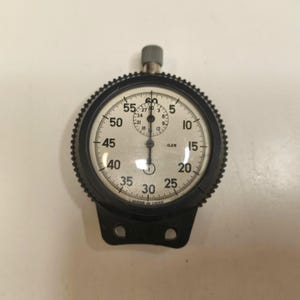 以下が含まれることがあります： 白い文字盤を持つ黒い機械式ストップウォッチ。外側の大きなダイヤルには5から60秒までの目盛りがあり、内側の小さなダイヤルには3から27までの目盛りがあります。文字盤には.025と印字されており、下端にはMade in USSRと記されています。