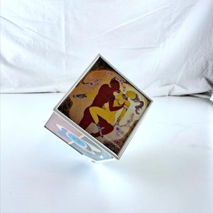 Peut inclure: Un cube encadré d'argent avec une peinture de deux figures enlacées, l'une rouge et l'autre jaune, sur un fond doré. Le cube est incliné, révélant un côté avec un motif abstrait coloré.