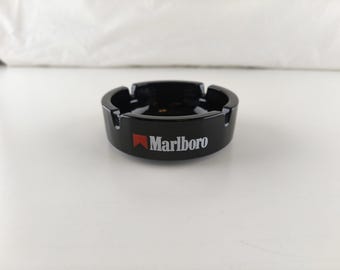 Könnte beinhalten: Ein runder, schwarzer Aschenbecher aus Keramik mit dem Marlboro-Logo, das in weißer Schrift mit einem roten Chevron-Symbol auf der Vorderseite aufgedruckt ist. Der Aschenbecher hat eine glatte, glänzende Oberfläche und vier gleichmäßige Kerben.
