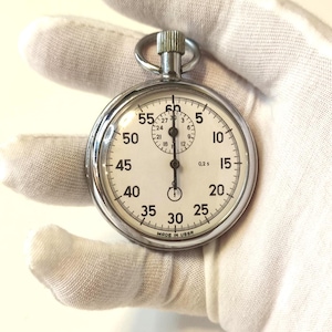 Puede incluir: Un cronómetro vintage con esfera blanca y números negros, sostenido por un guante blanco. El cronómetro tiene una carcasa plateada y un anillo en la parte superior. El texto "MADE IN USSR" es visible en la parte inferior.