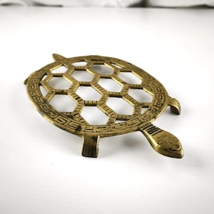 Puede incluir: Un salvamanteles de latón con forma de tortuga y diseño de panal en el centro. El salvamanteles tiene un borde decorativo y está diseñado para proteger las superficies de los utensilios de cocina calientes.