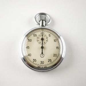 May include: A silver-toned mechanical stopwatch with a cream-colored face, black numerals, and a central winding crown with a ring. The dial features a main seconds track and a smaller sub-dial for minutes, with the text MADE IN USSR at the bottom.