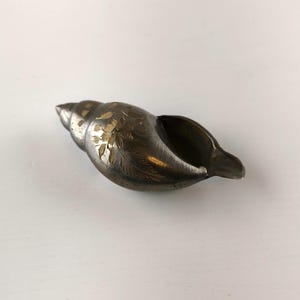 May include: A silver-toned seashell-shaped decorative object with etched floral designs. The shell has a dark interior and a pointed end. The etched designs are in a contrasting gold color, adding detail to the piece. The object is likely made of metal.