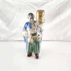 May include: A ceramic figurine of a person with a mustache wearing a blue coat and green trousers, holding a small barrel above their head. The barrel features a red logo and the text "СОЮЗ-ВИКТАН" in blue lettering. The figure has a white shirt.