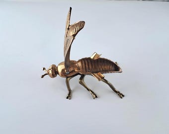 Könnte beinhalten: Eine Messing-Insektenskulptur mit detaillierten Flügeln und Beinen. Die Skulptur hat eine metallisch goldene Oberfläche und ist wie im Flug dargestellt. Der Körper des Insekts ist segmentiert, und die Flügel sind teilweise ausgebreitet.