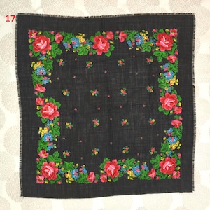 Op de afbeelding: Een vierkante zwarte sjaal met een bloemenpatroon. De sjaal heeft een rand van rode rozen, blauwe en gele bloemen en groene bladeren. De randen van de sjaal zijn gefranjerd.