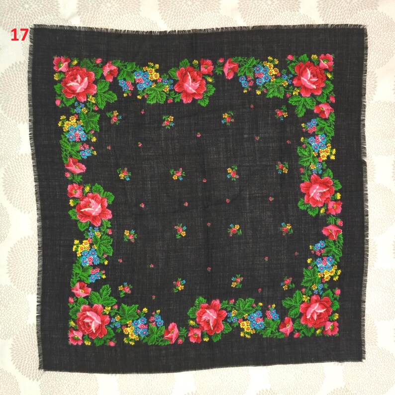 Op de afbeelding: Een vierkante zwarte sjaal met een bloemenpatroon. De sjaal heeft een rand van rode rozen, blauwe en gele bloemen en groene bladeren. De randen van de sjaal zijn gefranjerd.