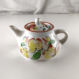 May include: White ceramic teapot with a lid, featuring hand-painted yellow, red, and green floral designs. The teapot has a curved spout and a rounded handle. The lid is topped with a small, white knob. The rim is trimmed with a brown border.