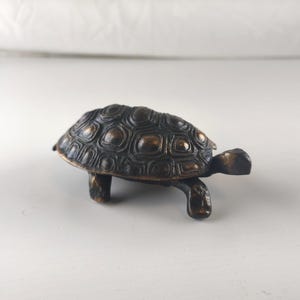Peut inclure: Une figurine de tortue de couleur bronze avec une carapace texturée et quatre pattes courtes. La tête et la queue de la tortue sont visibles, sur fond blanc. La sculpture est un objet décoratif.