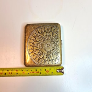 K&ouml;nnte beinhalten: Ein rechteckiges, antik-messingfarbenes Etui mit Kompassdesign und der Aufschrift "SAILING TO THE WORLD". Ein gelbes Ma&szlig;band ist neben dem Etui zu sehen. Das Etui ist ca. 9 cm lang.