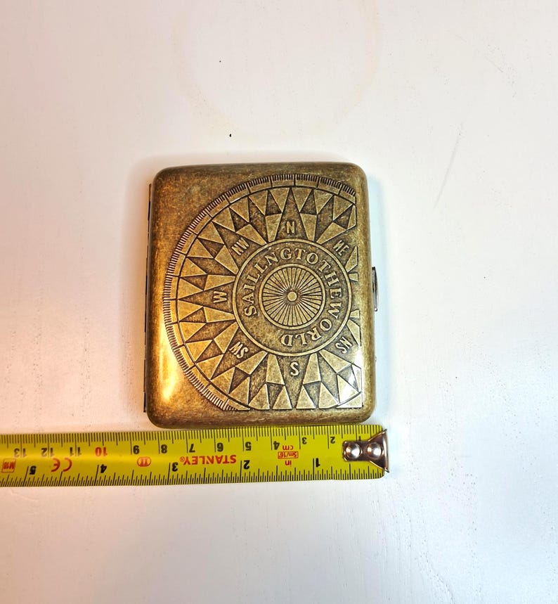K&ouml;nnte beinhalten: Ein rechteckiges, antik-messingfarbenes Etui mit Kompassdesign und der Aufschrift "SAILING TO THE WORLD". Ein gelbes Ma&szlig;band ist neben dem Etui zu sehen. Das Etui ist ca. 9 cm lang.