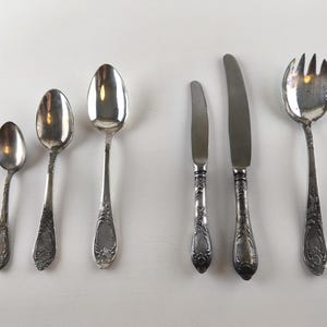 Op de afbeelding: Een verzameling antiek zilverwerk, waaronder lepels, messen en een serveervork, gerangschikt op een wit oppervlak. Het zilverwerk heeft sierlijke handvatten en een gepolijste afwerking. De set bevat verschillende maten lepels en messen.