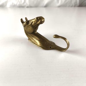 Op de afbeelding: Een messingkleurige paardenkophaak. De haak heeft de vorm van een paardenhoofd en -hals, met een gebogen haak die zich uitstrekt vanaf het lichaam. De haak is gemaakt van metaal en heeft een vintage uitstraling. De paardenkop is naar rechts gericht.