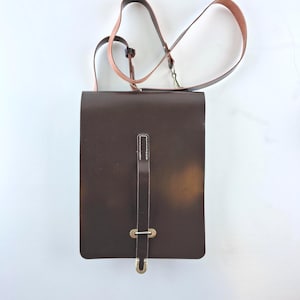 May include: A dark brown rectangular shoulder bag with a long strap. The bag has a flap closure with a vertical strap and a metal buckle. The shoulder strap is brown and pink.