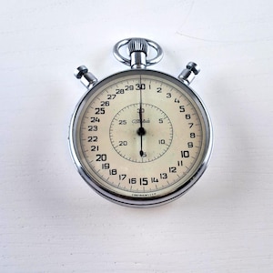 Puede incluir: Un cronómetro vintage con una caja plateada y una esfera color crema. El reloj presenta números y marcas en negro, con una esfera interna más pequeña. El cronómetro tiene dos botones y una corona de cuerda. Medidas en segundos.