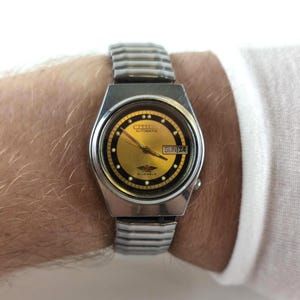 May include: A silver-tone Citizen automatic wristwatch featuring a round gold-colored dial with a black outer ring, luminous hour markers, and a day-date window at the three o'clock position. The watch includes a flexible metal expansion band.