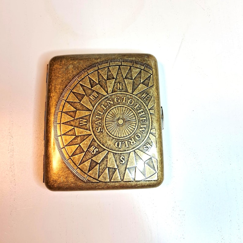 K&ouml;nnte beinhalten: Ein rechteckiges, antik-messingfarbenes Etui mit einem Kompassrosen-Design. Die Kompassrose ist in die Oberfl&auml;che ge&auml;tzt, mit den Worten "Sailing to the World", die die Mitte umkreisen. Das Etui hat einen Klappdeckel.
