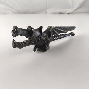 May include: A dark metal dragon figurine with open jaws, wings, and a detailed tail. The dragon is lying on a white surface. The figurine is likely a decorative item or a small sculpture.