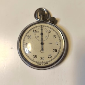 May include: A silver-toned mechanical stopwatch with a cream-colored face, black numerals, and two hands. The dial features a smaller sub-dial at the top, the text 0,2S, and the words MADE IN USSR printed at the bottom. It has a top crown and button.