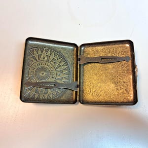 K&ouml;nnte beinhalten: Ge&ouml;ffnete antike Zigarettenetui aus Messing. Das Etui ist rechteckig mit einem Klappdeckel und Verschluss. Der Innenraum ist goldfarben, und der Deckel zeigt ein Kompassrosen-Design mit Richtungsmarkierungen. Das Etui ist ge&ouml;ffnet und gibt den Innenraum frei.