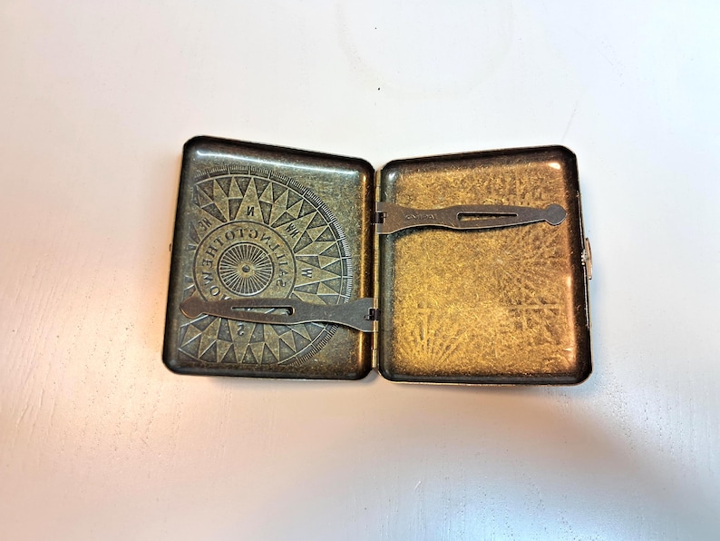 K&ouml;nnte beinhalten: Ge&ouml;ffnete antike Zigarettenetui aus Messing. Das Etui ist rechteckig mit einem Klappdeckel und Verschluss. Der Innenraum ist goldfarben, und der Deckel zeigt ein Kompassrosen-Design mit Richtungsmarkierungen. Das Etui ist ge&ouml;ffnet und gibt den Innenraum frei.