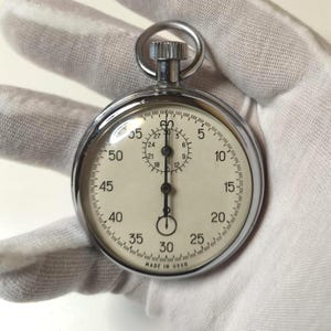 Puede incluir: Un cronómetro vintage con una carcasa plateada y una esfera blanca. La esfera presenta números y marcas negras para segundos y minutos. El texto "MADE IN USSR" es visible en la parte inferior. El cronómetro está sostenido por una mano enguantada.