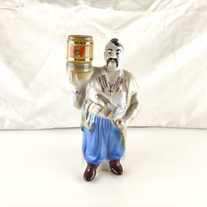 May include: A ceramic figurine of a person with a traditional moustache and hairstyle, wearing a white shirt with red and black embroidery, a striped sash, and blue trousers, holding a tan barrel with the text SV and SOY-VIKTAN printed on it.