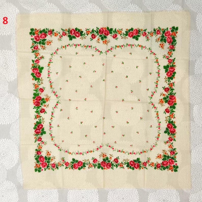 Op de afbeelding: Cr&egrave;mekleurig vierkant textiel met een bloemenontwerp. De rand heeft een guirlande van rode rozen, groene bladeren en oranje accenten. Het midden heeft een subtiel patroon van kleine bloemmotieven.