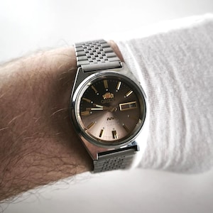 May include: A silver-toned Orient watch with a brown and gold face. The watch has a date display and a metal band. The watch is on a wrist, with a white sleeve in the background.