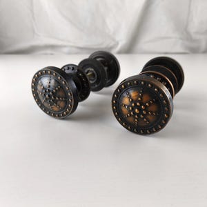 Puede incluir: Dos tiradores de puerta antiguos de bronce. Cada tirador presenta un diseño circular con un acabado en bronce oscuro y pequeños puntos en relieve. Los tiradores se exhiben sobre una superficie blanca.