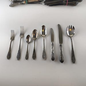 Op de afbeelding: Een verzameling antiek zilverwerk, waaronder vorken, lepels en messen, gerangschikt op een wit oppervlak. Het zilverwerk heeft sierlijke, gedetailleerde handvatten en een aangeslagen zilveren afwerking, wat duidt op leeftijd en gebruik. De set bevat vorken, lepels en messen in verschillende maten.