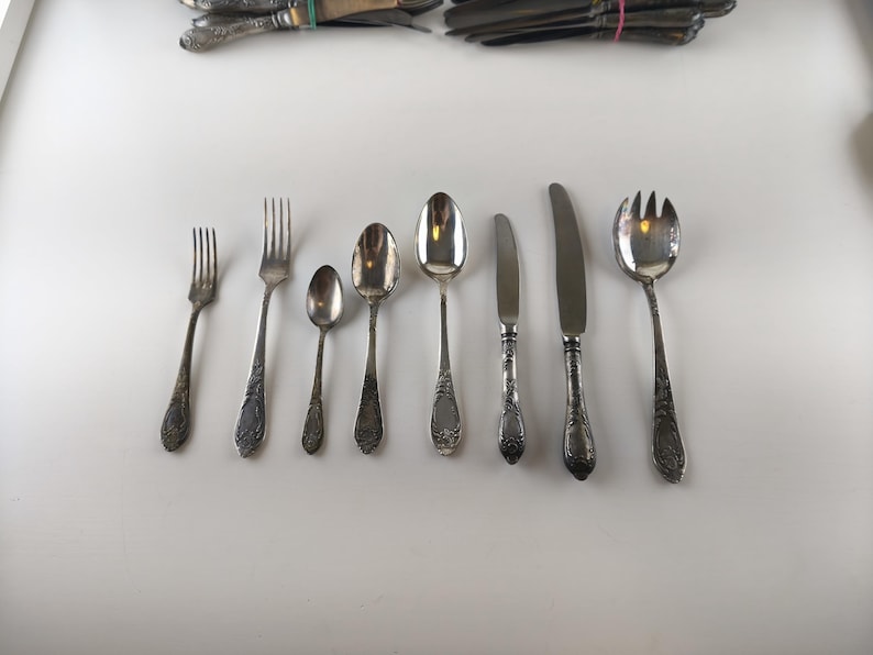 Op de afbeelding: Een verzameling antiek zilverwerk, waaronder vorken, lepels en messen, gerangschikt op een wit oppervlak. Het zilverwerk heeft sierlijke, gedetailleerde handvatten en een aangeslagen zilveren afwerking, wat duidt op leeftijd en gebruik. De set bevat vorken, lepels en messen in verschillende maten.