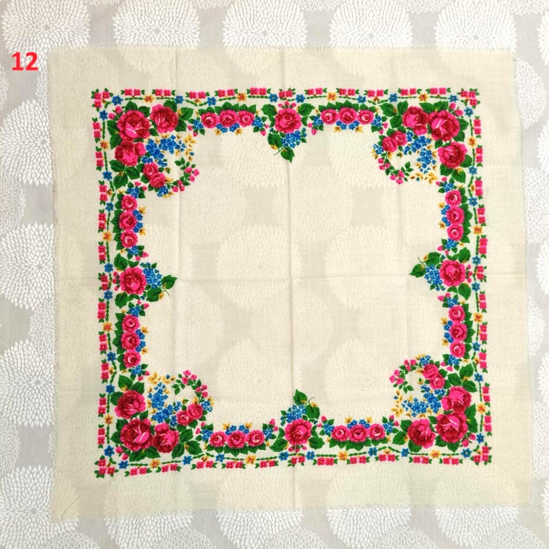 May include: Cream-colored square scarf with a floral border. The border features pink roses, blue and yellow flowers, and green leaves. The number "12" is in the upper left corner. The scarf is likely made of a soft fabric.