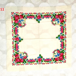 May include: Square, off-white scarf with a floral border. The border features pink roses, blue flowers, and green leaves. The scarf is made of a lightweight fabric.