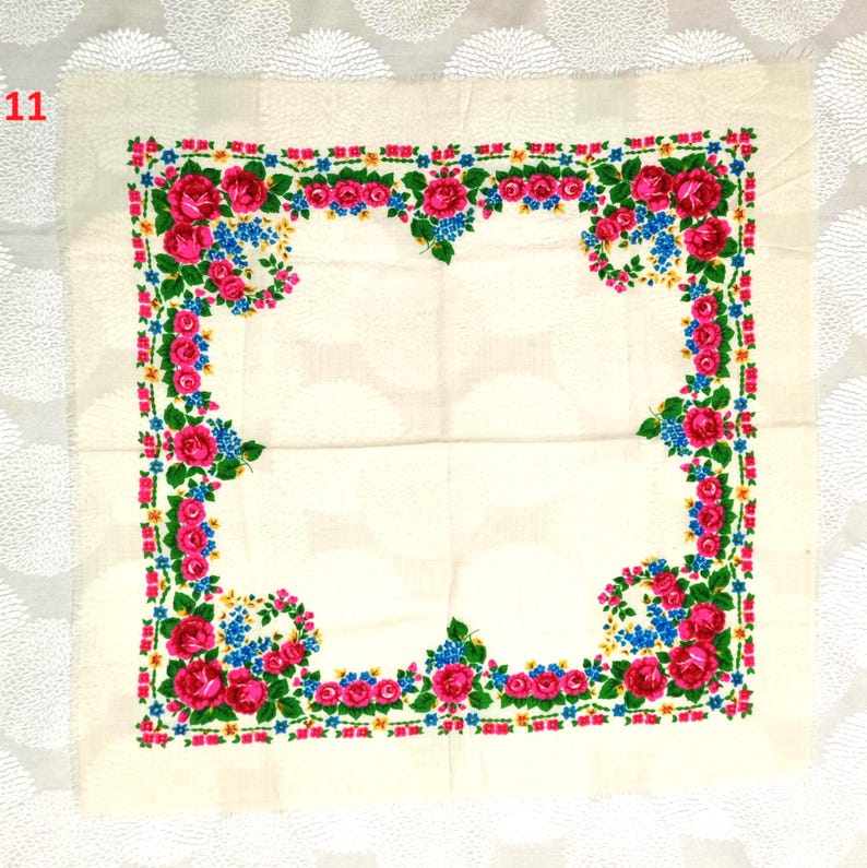 May include: Square, off-white scarf with a floral border. The border features pink roses, blue flowers, and green leaves. The scarf is made of a lightweight fabric.