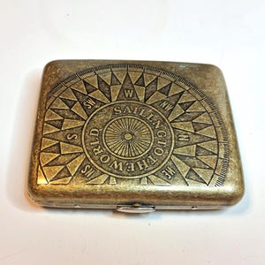 K&ouml;nnte beinhalten: Ein rechteckiges Metallgeh&auml;use im antiken Stil mit einem Kompassrosen-Design. Die Kompassrose ist in die Oberfl&auml;che ge&auml;tzt und tr&auml;gt die Worte "SAILNG TOTHEWORLD". Das Geh&auml;use hat einen Klappverschluss und eine angelaufene Bronzeoberfl&auml;che.