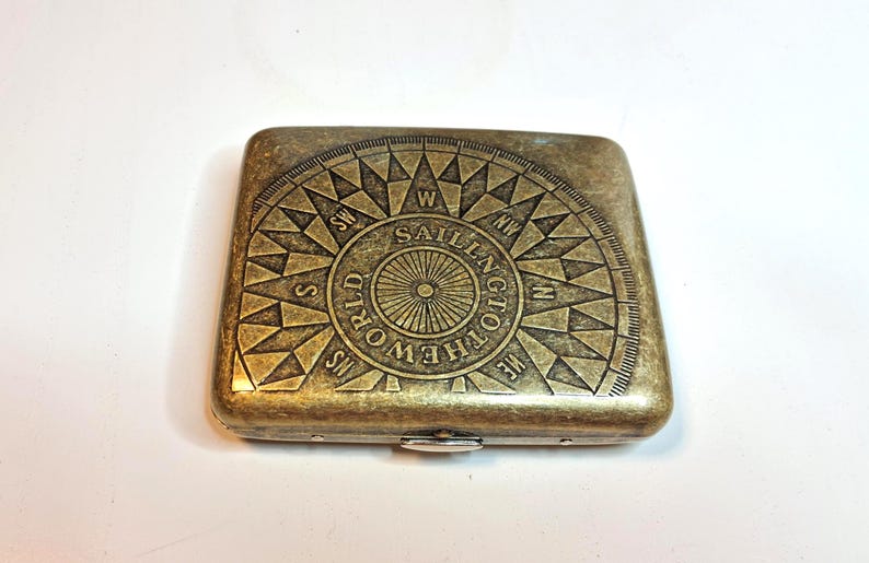 K&ouml;nnte beinhalten: Ein rechteckiges Metallgeh&auml;use im antiken Stil mit einem Kompassrosen-Design. Die Kompassrose ist in die Oberfl&auml;che ge&auml;tzt und tr&auml;gt die Worte "SAILNG TOTHEWORLD". Das Geh&auml;use hat einen Klappverschluss und eine angelaufene Bronzeoberfl&auml;che.