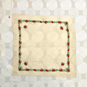 Op de afbeelding: Cr&egrave;mekleurig vierkant textiel met een bloemenrand. De rand heeft rode rozen, groene bladeren en kleine blauwe accenten. Vier rozenclusters zijn geplaatst in de hoeken en het midden van het textiel.