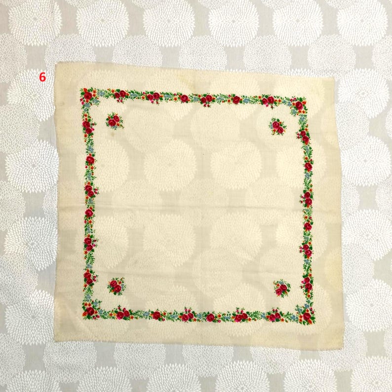 Op de afbeelding: Cr&egrave;mekleurig vierkant textiel met een bloemenrand. De rand heeft rode rozen, groene bladeren en kleine blauwe accenten. Vier rozenclusters zijn geplaatst in de hoeken en het midden van het textiel.