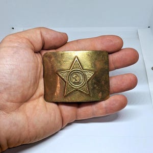 Könnte beinhalten: Rechteckige Messing-Gürtelschnalle mit erhabenem Sternemblem und Hammer-und-Sichel-Symbol. Die Schnalle hat ein angelaufenes, gealtertes Aussehen. Ungefähre Abmessungen: 8 cm x 4 cm.